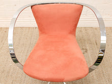 Load image into Gallery viewer, A mid-century modern accent chair with a curved, polished chrome tubular frame and a coral pink suede or velvet upholstered seat and backrest, set on a woven jute rug.
