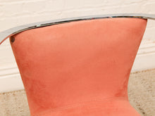 Load image into Gallery viewer, Close-up of a coral pink suede chair back with a curved chrome metal rim, set against a white brick wall and beige carpet.
