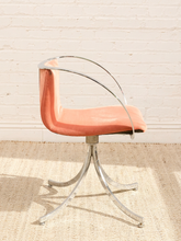 Load image into Gallery viewer, A mid-century modern accent chair with a peach-colored velvet seat and backrest, supported by a chrome swivel base and curved chrome armrests, set against a white brick wall and light beige rug.
