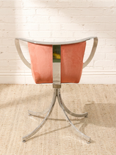Load image into Gallery viewer, A modern accent chair with a curved backrest upholstered in a terracotta pink fabric, a central chrome column, and curved chrome side arms and legs, set against a white brick wall and beige jute rug.
