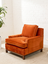 Load image into Gallery viewer, A modern orange velvet armchair with a matching back cushion, set on a light beige carpet against a white brick wall background.
