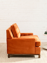 Load image into Gallery viewer, A modern orange velvet armchair with a low wooden base, positioned against a white brick wall background.
