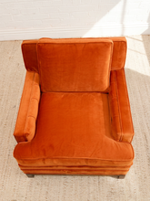Load image into Gallery viewer, An orange velvet armchair with a high backrest, photographed from a top-down angle on a textured beige rug against a white brick wall.
