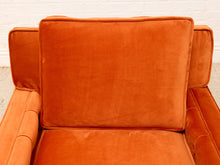 Load image into Gallery viewer, Close-up of an orange velvet armchair with a large back cushion and seat cushion, set against a white brick wall background.
