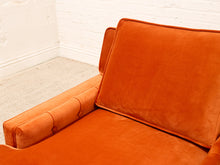 Load image into Gallery viewer, A close-up of a modern orange velvet armchair in a room corner with a white brick wall and light carpet.
