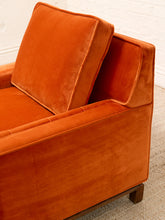 Load image into Gallery viewer, Close-up of a modern armchair upholstered in rich burnt orange velvet, showing the back cushion, armrest, and corner detail against a white brick wall and light carpet.
