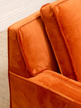 Load image into Gallery viewer, Close-up of an orange velvet armchair corner with thick padded cushions and clean lines, set against a light neutral wall and carpet.
