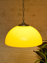 Load image into Gallery viewer, A glowing yellow dome-shaped pendant light with a dark metal top and chain, hanging against a warm brick wall with a plant leaf visible in the lower right corner.
