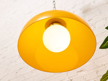Load image into Gallery viewer, A modern pendant light with a wide, shallow yellow-orange dome shade and a glowing white globe bulb, hanging against a white brick wall.
