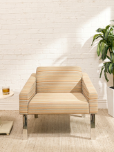 Load image into Gallery viewer, A modern armchair with beige and orange striped upholstery and chrome legs, set against a white brick wall with a potted plant and a small side table nearby.
