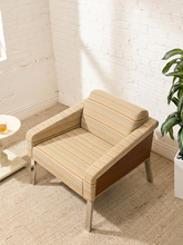 Load image into Gallery viewer, A mid-century modern accent chair with a beige and brown striped fabric upholstery and a dark wood side panel, set in a bright room with a white brick wall, beige carpet, and a potted plant.
