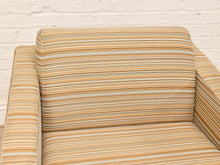 Load image into Gallery viewer, A close-up of a striped upholstered armchair with a backrest and armrests, featuring horizontal stripes in beige, mustard yellow, and brown tones, set against a white brick wall.
