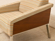 Load image into Gallery viewer, A mid-century modern armchair featuring a warm brown wood veneer side panel and striped upholstery in beige, cream, and orange tones, shown on a textured rug.
