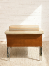 Load image into Gallery viewer, A mid-century modern wooden bench with a striped cushion on the seat and chrome legs, set against a white brick wall with a diagonal patch of sunlight.
