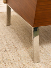Load image into Gallery viewer, Close-up of a mid-century modern wooden sideboard or cabinet with polished chrome legs, resting on a textured beige woven rug.
