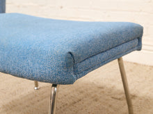 Load image into Gallery viewer, A close-up of a mid-century modern upholstered stool with a textured blue fabric seat cushion and chrome legs, set against a light brick wall and beige carpet.
