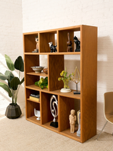 Load image into Gallery viewer, A tall, multi-compartment wooden bookcase with a warm medium-brown finish, styled with various decorative objects including figurines, a bowl, a vase of yellow flowers, and a geometric sculpture, set against a white brick wall.

