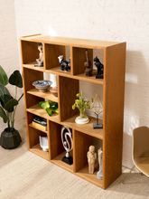 Load image into Gallery viewer, A tall wooden open shelving unit with multiple cubby compartments, styled with various decorative objects, set against a white brick wall in a room.
