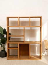 Load image into Gallery viewer, A light natural wood modular bookcase with multiple open shelves, set against a white brick wall with a potted plant on the left and a chair on the right.
