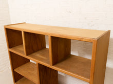 Load image into Gallery viewer, A mid-century modern wooden bookcase with six cubby shelves, made of warm medium-brown wood, set against a white brick wall.
