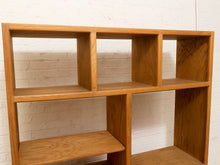 Load image into Gallery viewer, A mid-century modern wooden shelving unit with a warm medium-brown finish, featuring a grid of open cubbies against a white painted brick wall.
