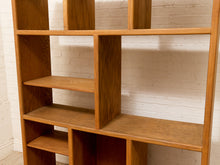 Load image into Gallery viewer, A mid-century modern wooden bookcase with a warm honey finish, featuring open cubby shelves, set against a white brick wall.
