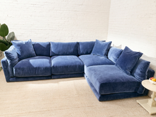 Load image into Gallery viewer, A large L-shaped sectional sofa upholstered in textured blue velvet, styled in a bright living room with a white brick wall and light beige rug.

