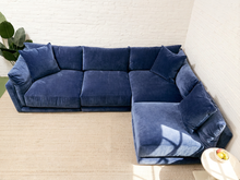 Load image into Gallery viewer, A large L-shaped sectional sofa upholstered in deep navy blue velvet, positioned in a bright room with a white brick wall and a light beige rug.
