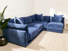 Load image into Gallery viewer, A modern sectional sofa upholstered in deep blue velvet, styled in a minimalist room with a white brick wall, beige carpet, and a potted plant on the left.
