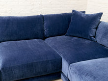 Load image into Gallery viewer, A modern sectional sofa upholstered in deep navy blue velvet fabric, featuring a matching throw pillow, set against a white brick wall background.
