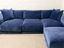 Load image into Gallery viewer, A large L-shaped sectional sofa upholstered in deep blue velvet fabric, shown in a living room setting with a light beige rug and a white brick wall background.

