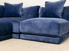 Load image into Gallery viewer, A close-up of a plush sectional sofa upholstered in deep navy blue velvet, featuring thick seat cushions, back cushions, and matching throw pillows, set against a white brick wall and light beige carpet.
