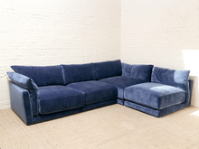 Load image into Gallery viewer, A modern L-shaped sectional sofa upholstered in deep navy blue velvet, positioned in a minimalist room with a white brick wall and light beige carpet.
