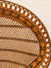 Load image into Gallery viewer, Close-up of a woven rattan chair back showing the curved orange-brown cane weave with black accent stripes and a decorative looped border, set against a white brick wall background.
