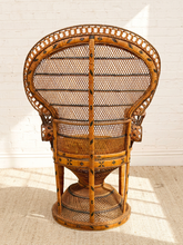 Load image into Gallery viewer, A tall, woven rattan accent chair with a high back, turned toward the back to show the detailed weave and carved frame, set against a white brick wall.
