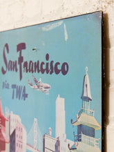 Load image into Gallery viewer, A vintage-style San Francisco travel sign with a light blue background, featuring the text 'San Francisco via TWA' and an illustration of the city skyline, including the Golden Gate Bridge and a tower, mounted against a white brick wall.

