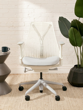 Load image into Gallery viewer, A modern white mesh office chair with a white padded seat and a five-star wheeled base, styled in a bright room with a white brick wall, a potted plant, and a side table.

