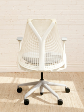 Load image into Gallery viewer, A modern white mesh office chair with a five-star wheeled base, positioned against a white brick wall on a textured beige rug.
