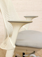Load image into Gallery viewer, Close-up of a modern ergonomic office chair with a white mesh back, sculptural white armrests, and a light gray upholstered seat cushion, set against a white brick wall.
