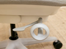 Load image into Gallery viewer, Close-up of the base mechanism of an office chair, showing a white plastic lever, a circular ring handle, and a black caster wheel on a beige carpeted floor.
