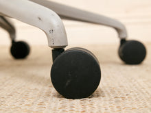 Load image into Gallery viewer, Close-up of a black caster wheel attached to a light gray metal chair arm, resting on a beige woven rug.

