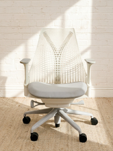 Load image into Gallery viewer, A modern office chair with a white mesh backrest, gray seat cushion, and light gray five-star base with black casters, set against a white brick wall with soft window shadows.
