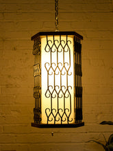 Load image into Gallery viewer, A hanging rectangular pendant light with a glowing warm white frosted center panel and dark metal scrollwork side guards, suspended against a yellow-lit brick wall.
