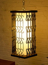 Load image into Gallery viewer, A hanging rectangular metal lantern light with a glowing warm white center panel and dark wrought iron scrollwork, suspended against a white brick wall.
