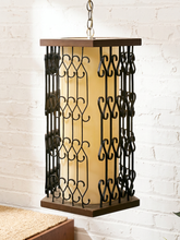 Load image into Gallery viewer, A hanging lantern-style pendant light with a warm cream glass shade and dark metal scrollwork guards, suspended against a white painted brick wall.
