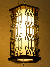 Load image into Gallery viewer, A hanging lantern-style light fixture with a glowing warm white center panel and dark metal scrollwork sides, shown against a textured white wall.
