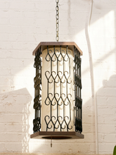 Load image into Gallery viewer, A hanging lantern-style pendant light with a cream cylindrical shade and dark metal scrollwork frame, suspended by a chain against a white painted brick wall.
