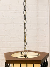 Load image into Gallery viewer, A close-up of a hanging lantern light fixture with a hexagonal dark brown wood top, black metal frame, and a warm cream-colored glass shade, suspended by a dark metal chain against a white brick wall.
