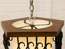 Load image into Gallery viewer, Hexagonal pendant light fixture with a dark brown wood top frame, black metal scrollwork, and a warm cream-colored glass shade, shown hanging against a white brick wall.
