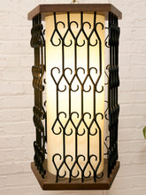 Load image into Gallery viewer, A rectangular wall sconce with a glowing warm white cylindrical shade, overlaid by black wrought iron scrollwork, set in a dark brown wooden frame against a white brick wall.
